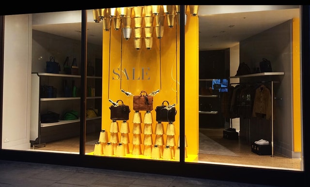 Retail store window display featuring suspended handbags and stacked gold cone props against a yellow backdrop with a prominent sale graphic, viewed through the storefront glazing.