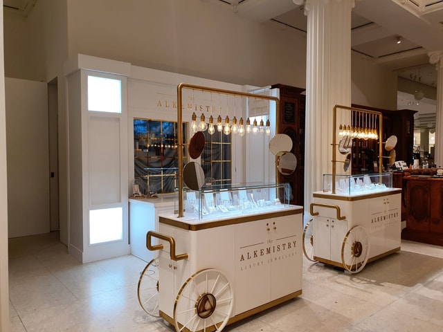 White and gold mobile jewellery display carts with glass cases and pendant lights branded 'The Alkemistry' in a polished retail store setting.