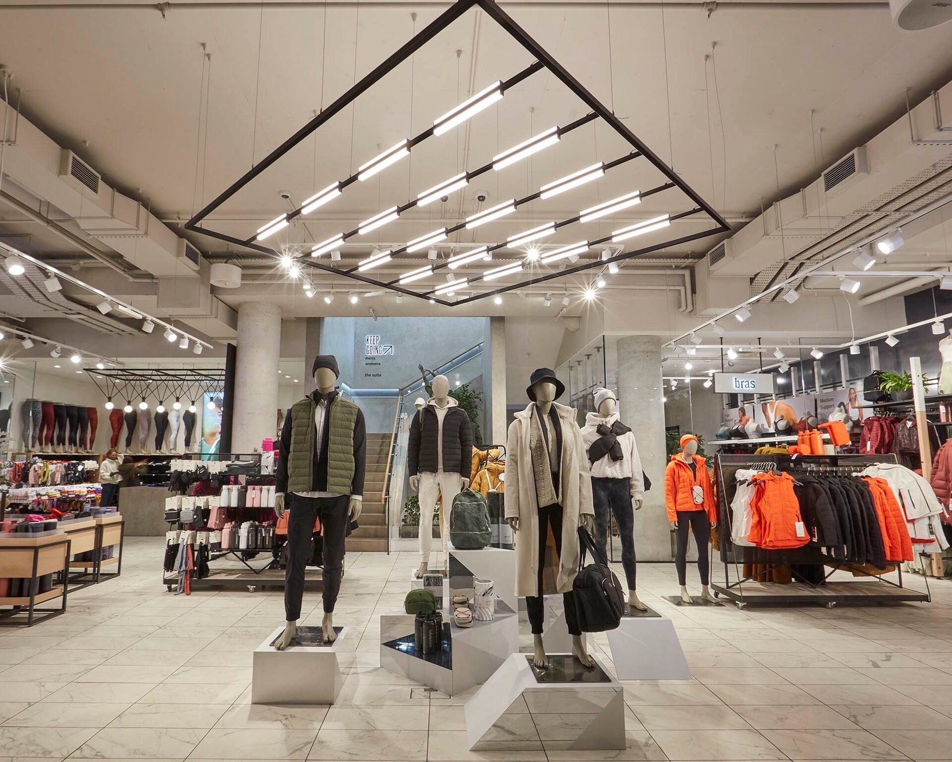 Bright retail clothing store interior with mannequins on display plinths, clothing racks and a visible 'bras' department sign; modern lighting and tiled flooring in a clean store environment.