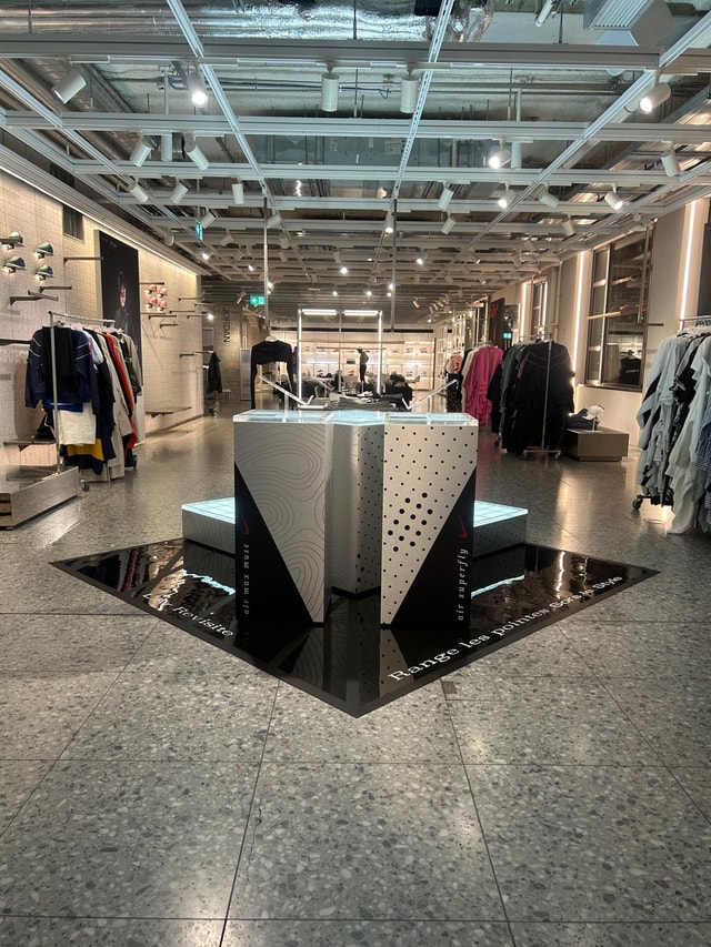 Interior retail store with a central branded product display island, clothing racks and printed floor graphics in a well-lit Nike shop environment.
