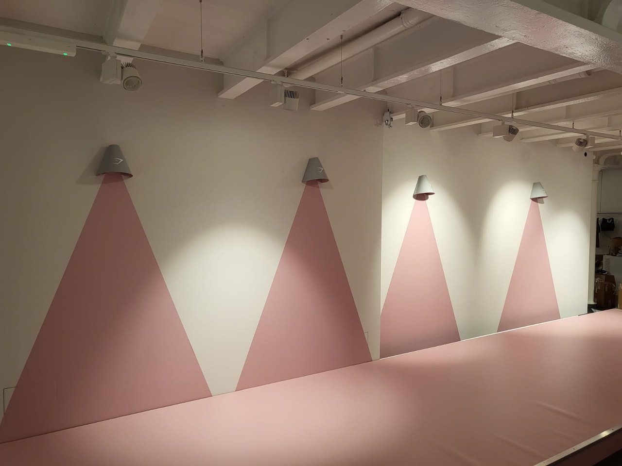 Minimal retail interior with repeated pink triangular wall graphics lit by cone-shaped wall lights and a long pink display table, showcasing a designed shop fit-out.