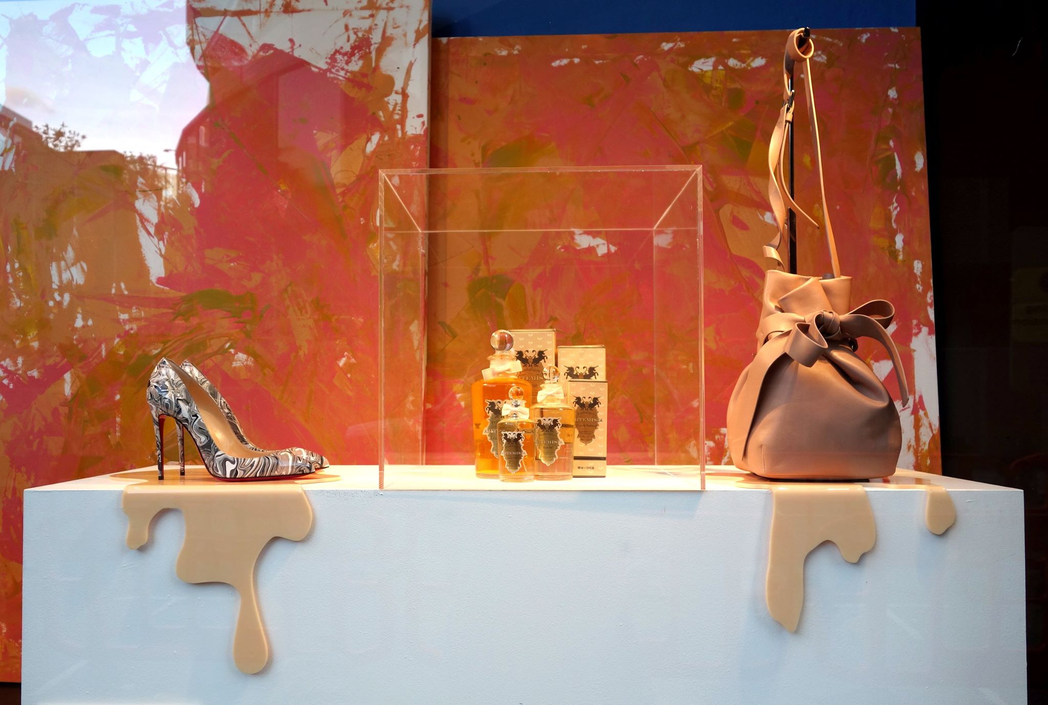 Stylish shop window display featuring a high-heeled shoe, leather handbag and perfume bottles showcased in a clear acrylic cube against a colourful painted backdrop.
