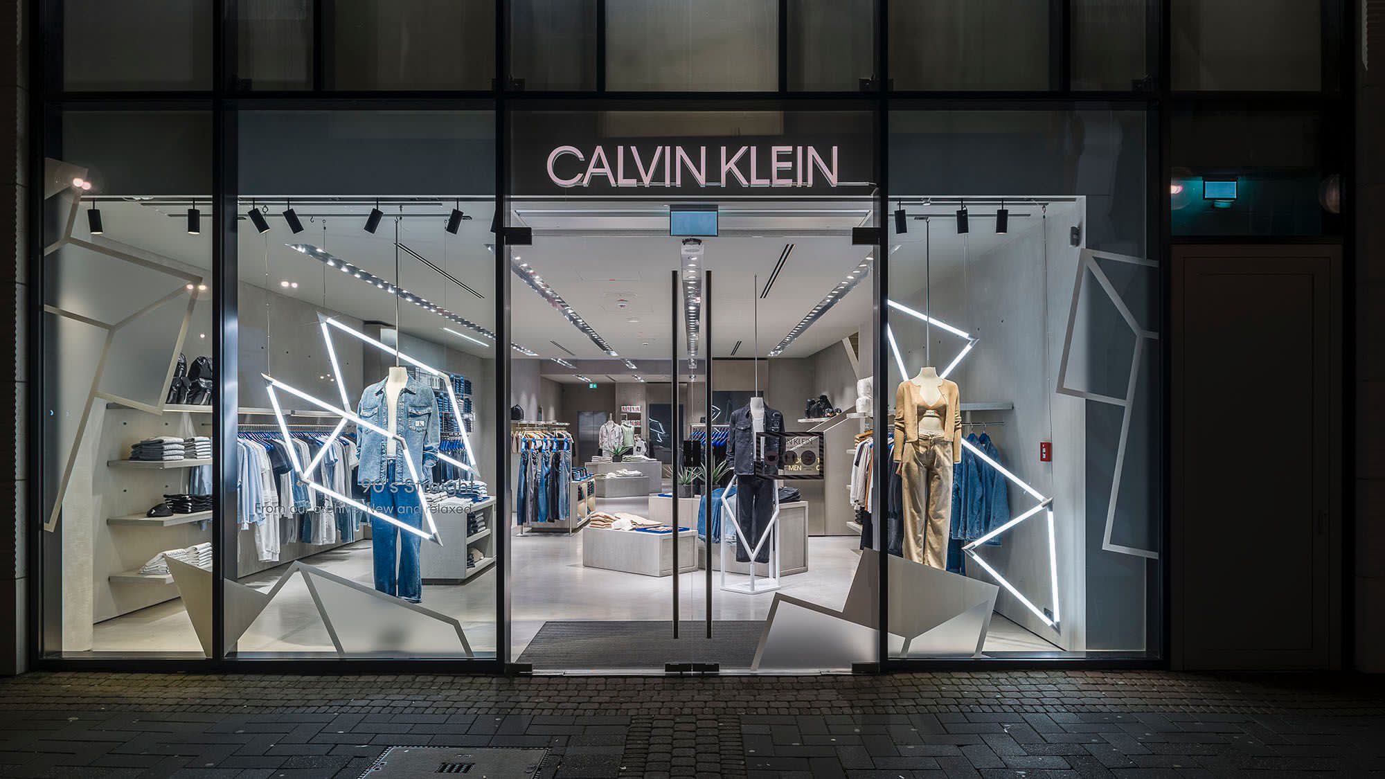 Calvin Klein storefront at night featuring an illuminated brand fascia, large glazed windows with geometric window graphics, mannequins and premium interior retail displays.
