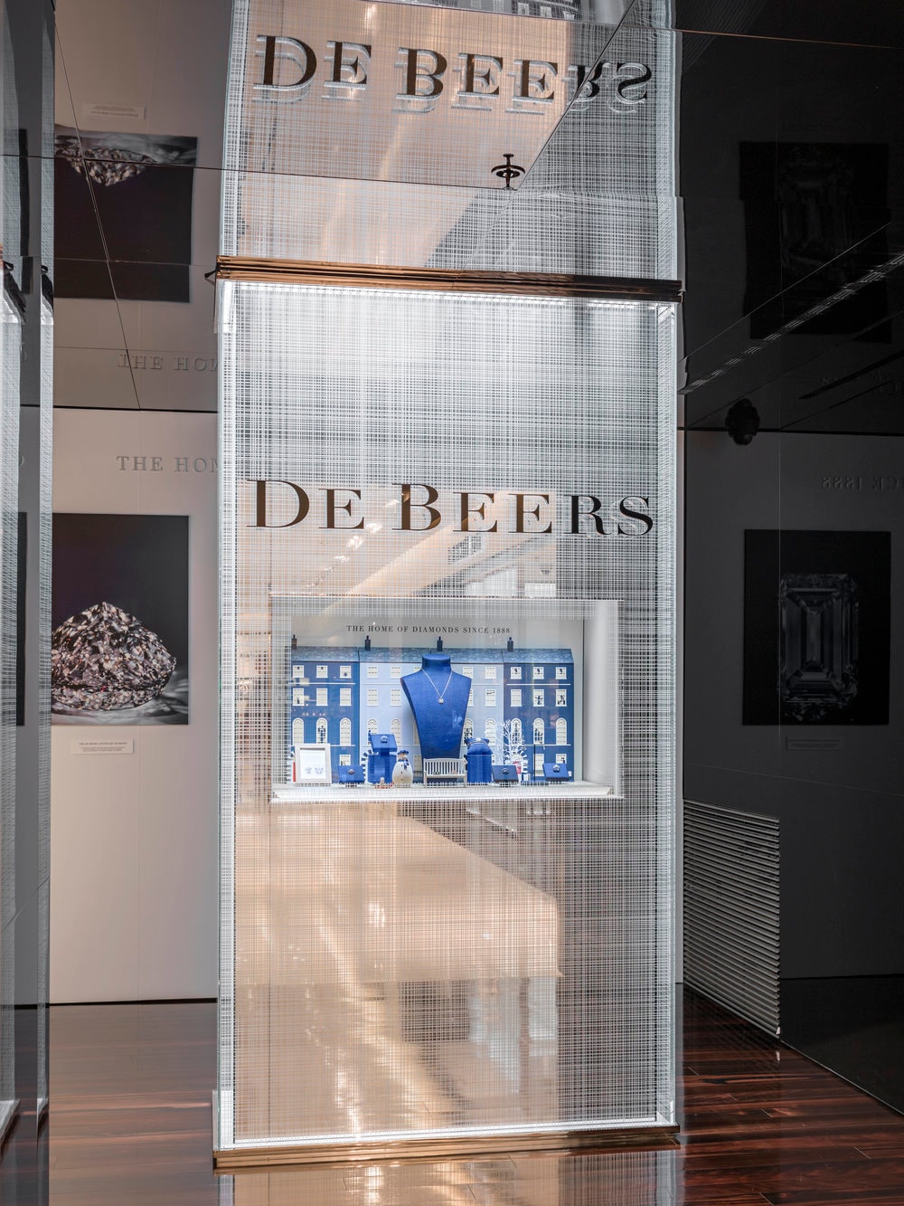 De Beers shopfront with glass-mounted brand signage and a curated jewellery window display behind a textured glass panel in a luxury retail interior.