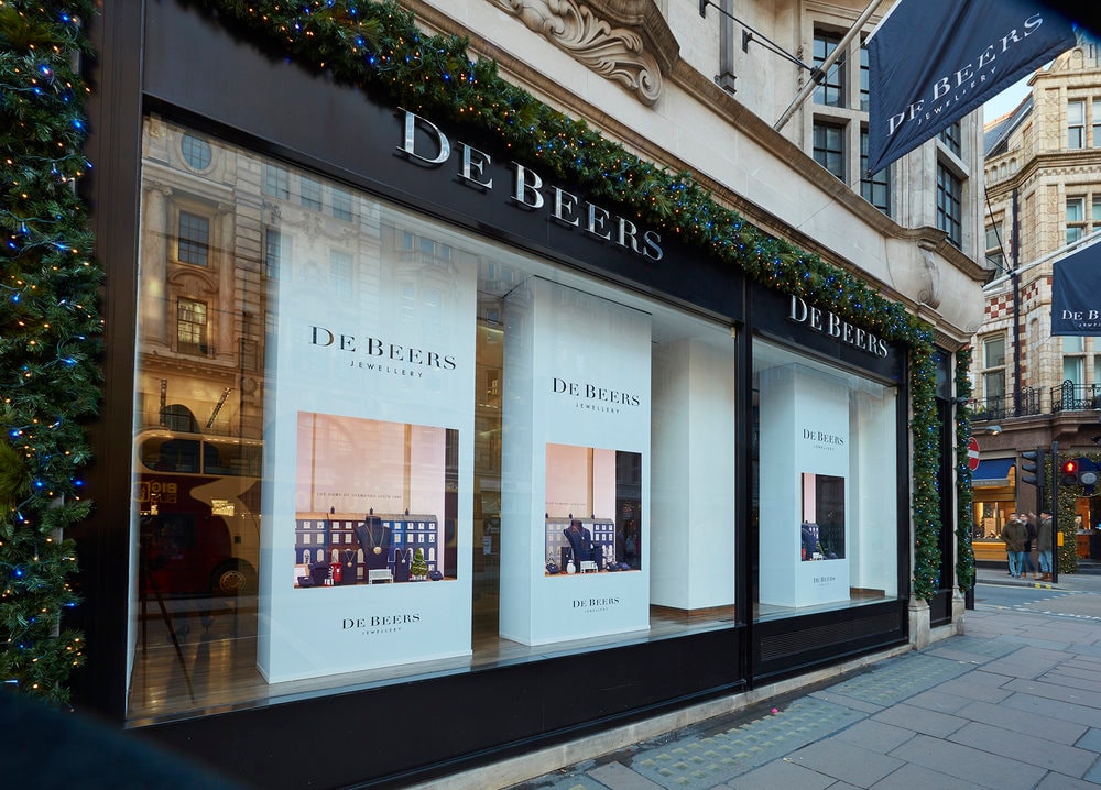 De Beers jewellery storefront with raised fascia lettering and retail window displays framed by festive garlands on a London street. High-quality exterior shot showing brand signage and window manifestations.