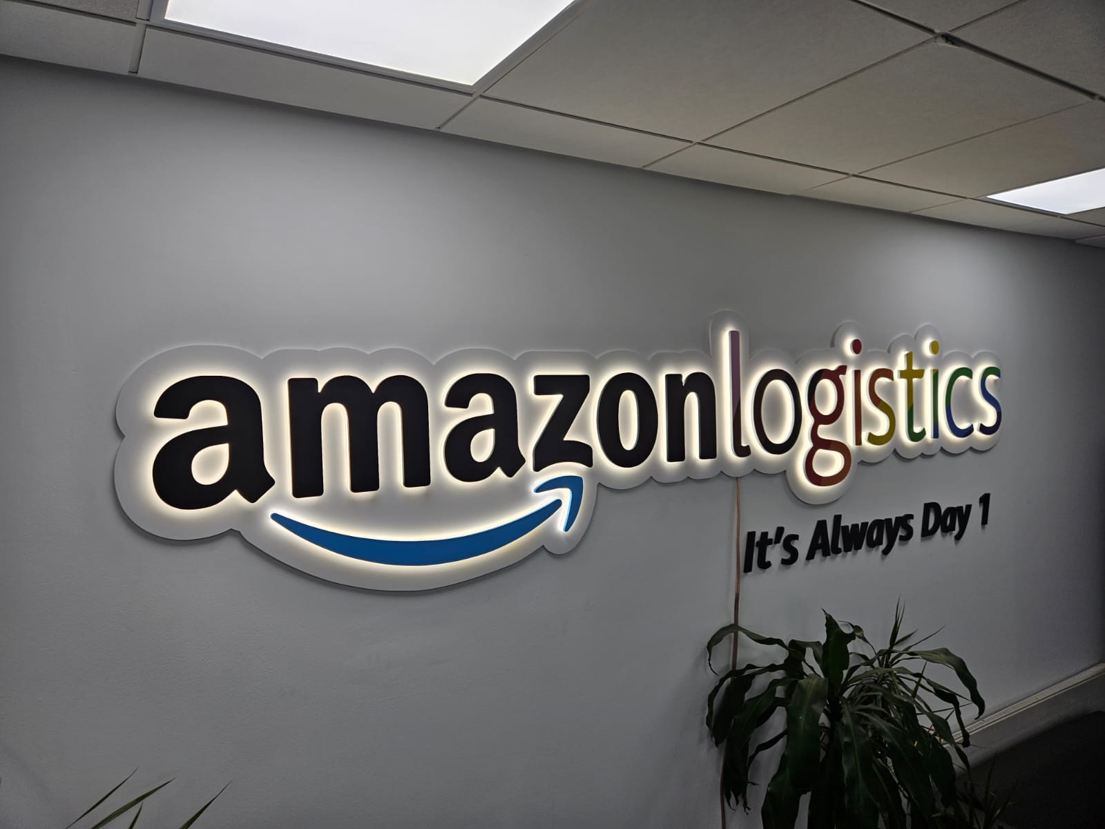 Edge-lit Amazon Logistics wall sign with halo LED backlighting installed in an office reception, featuring the company smile logo and the slogan 'It's Always Day 1'.