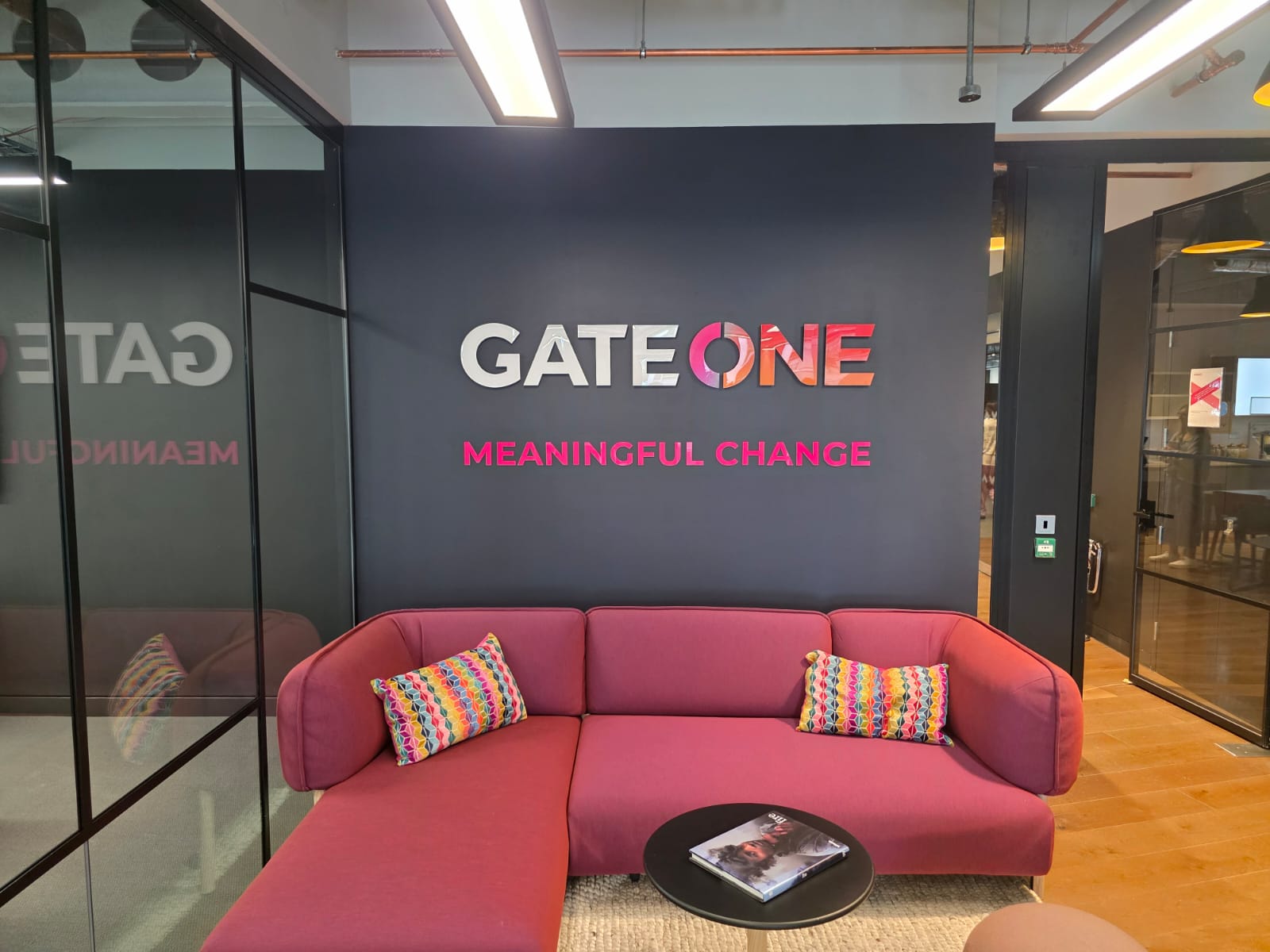 Office reception with installed 3D 'GATE ONE' wall letters and 'MEANINGFUL CHANGE' tagline above a pink sofa in a modern workspace.