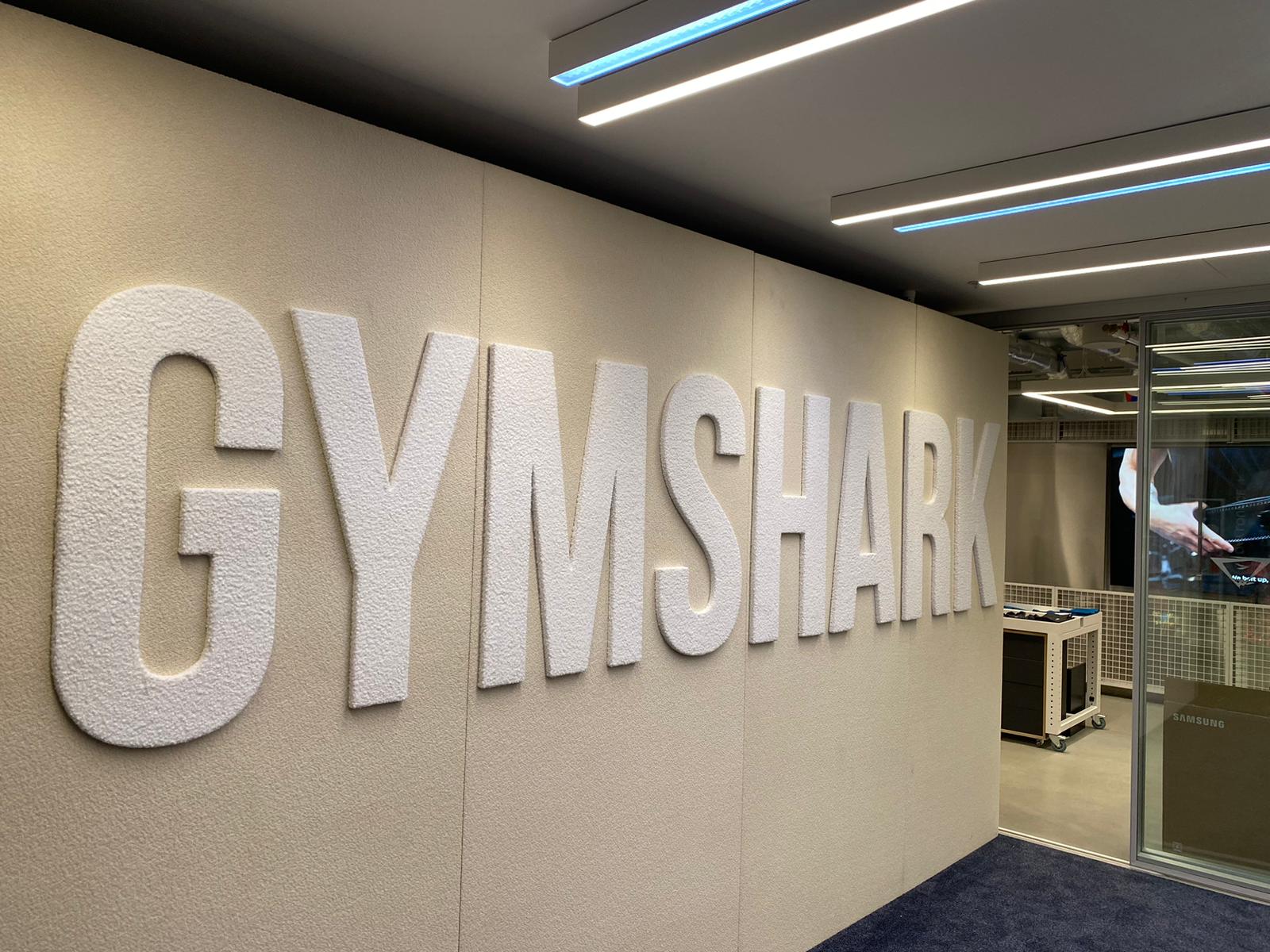 Large raised textured white letters spelling GYMSHARK mounted on an interior fabric wall in an office/reception area, with a glass meeting room and linear LED lighting.