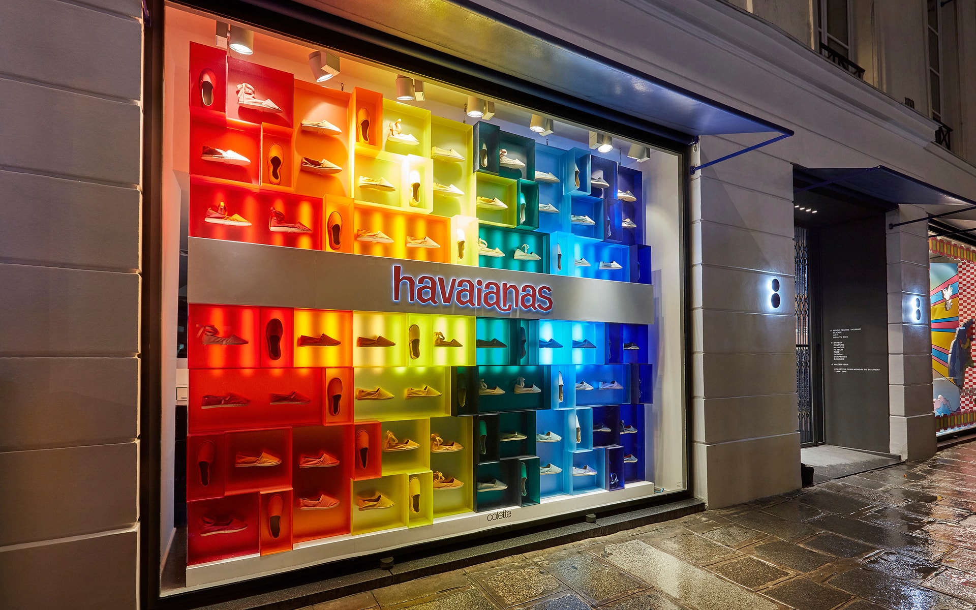 Bright Havaianas storefront window with rainbow-coloured cubby product display and illuminated brand signage, photographed from the street at night.