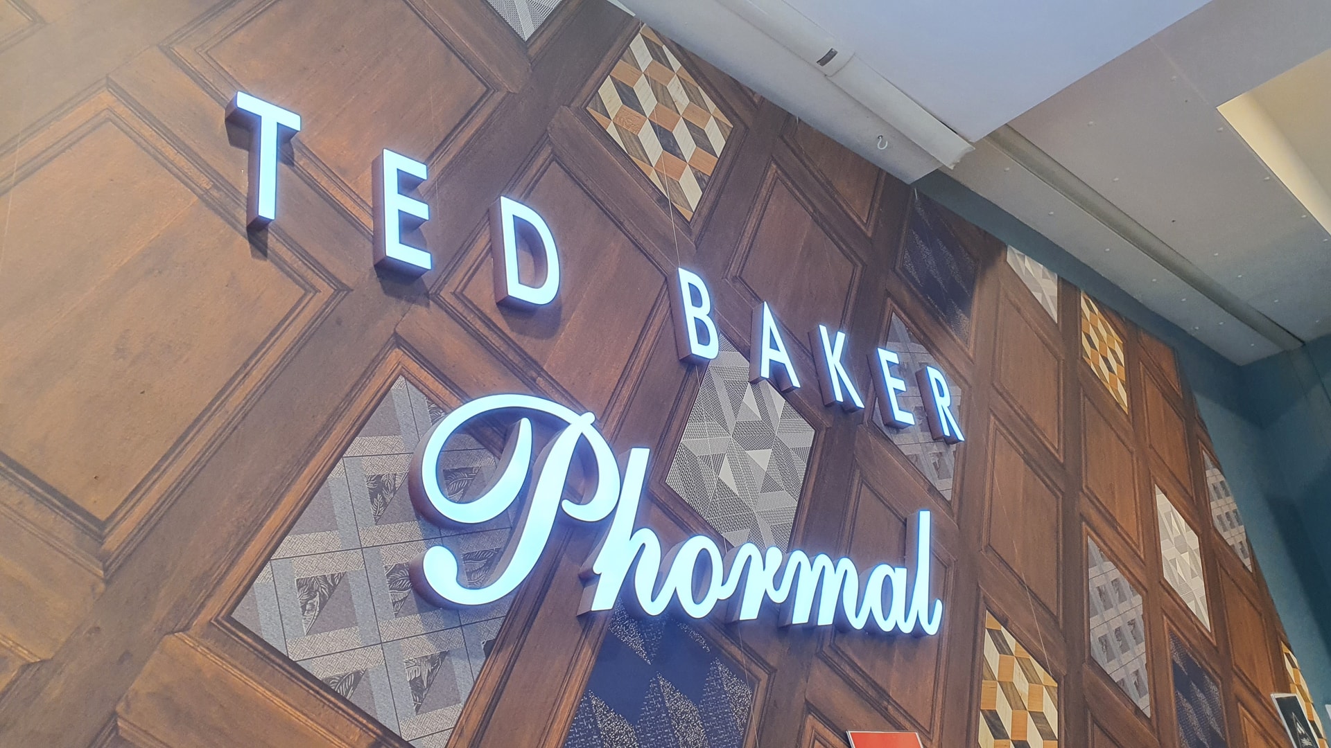 Illuminated halo-lit built-up letters spelling 'Ted Baker Phormal' mounted on a decorative timber feature wall inside a retail store.