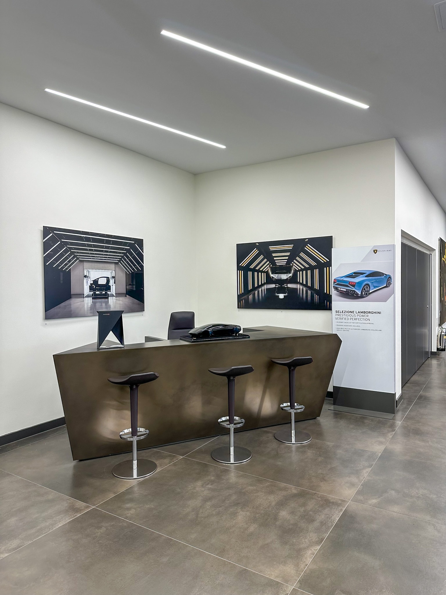 Modern Lamborghini showroom reception featuring a branded freestanding display and sleek reception desk with bar stools, set against framed automotive photography and minimalist lighting.