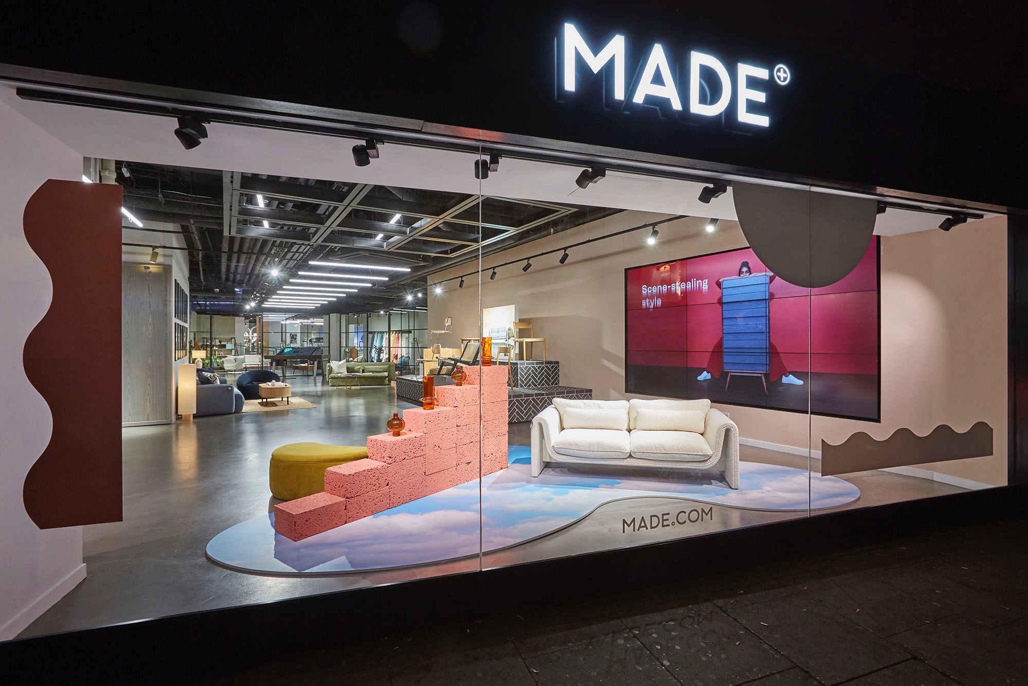 Illuminated MADE storefront sign above a retail window display featuring a sofa and staged showroom interior with a video wall.