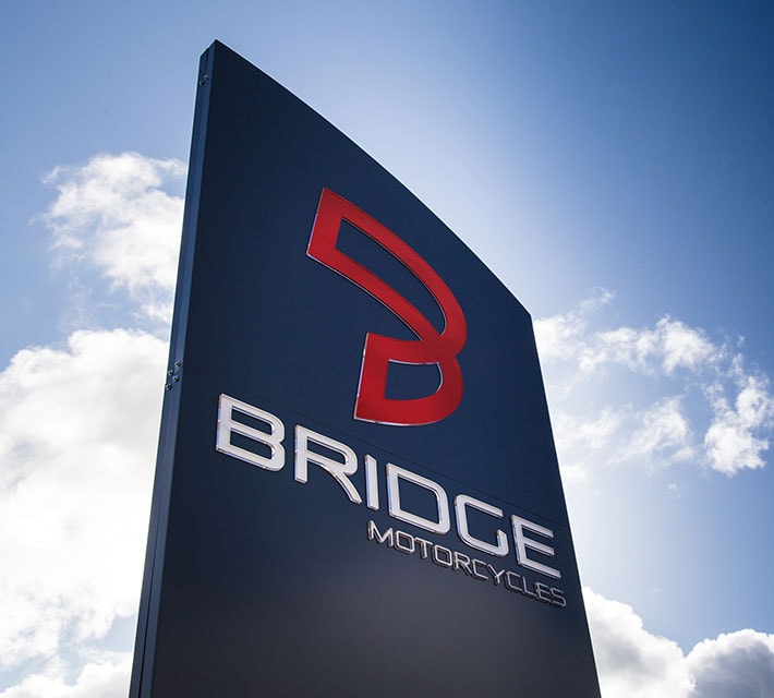 Exterior freestanding sign for Bridge Motorcycles featuring raised chrome letters and a red logo on a curved dark panel against a blue sky.