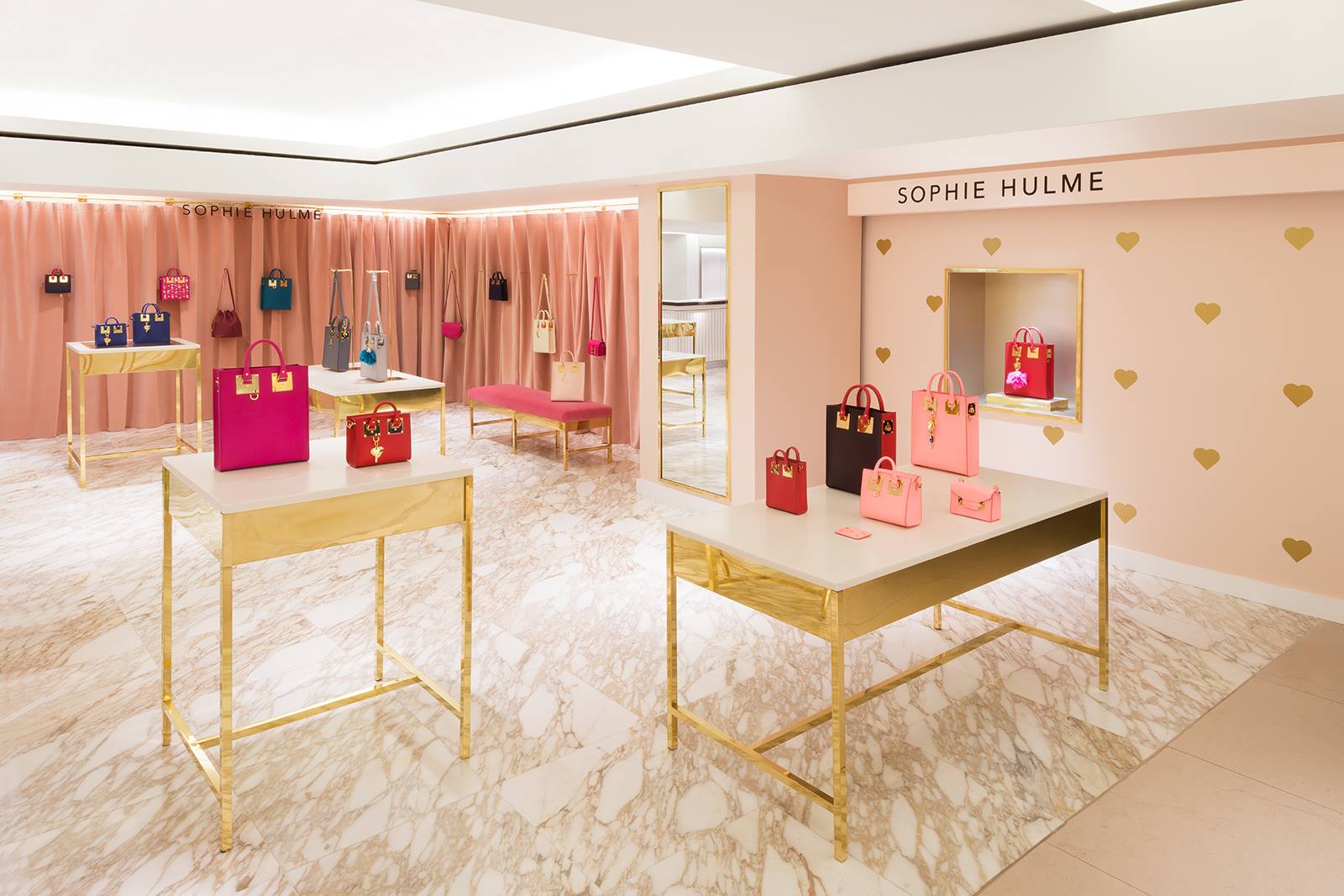 Luxury Sophie Hulme boutique interior with gold display tables, pink feature walls and branded signage showcasing handbags in a high-end retail setting.