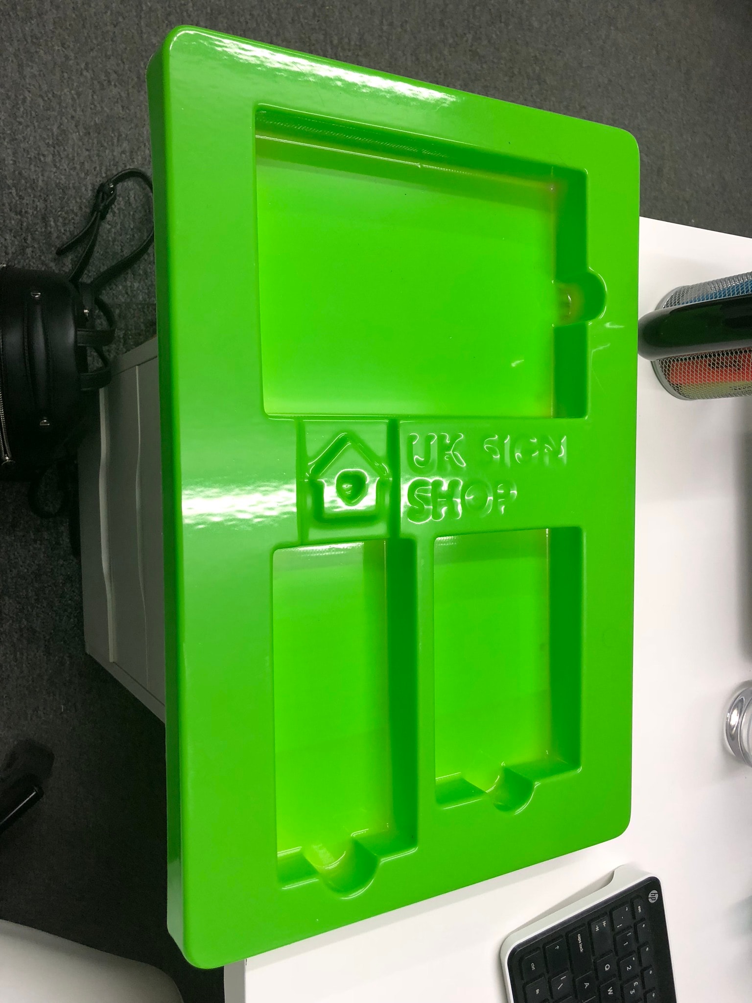 Glossy green vacuum-formed plastic tray with recessed compartments and an embossed 'UK SIGN SHOP' logo, photographed on a desk. Example of a thermoformed packaging or display insert showing brand embossing.