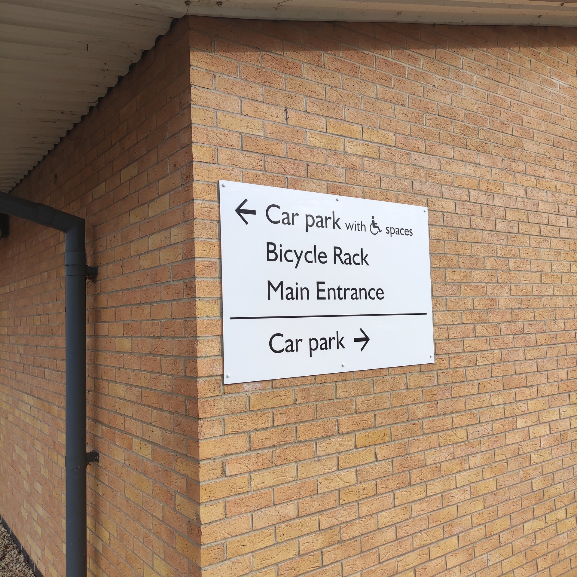 Exterior directional wayfinding sign mounted to a brick wall indicating car park, bicycle rack and main entrance with arrows and accessibility symbol.