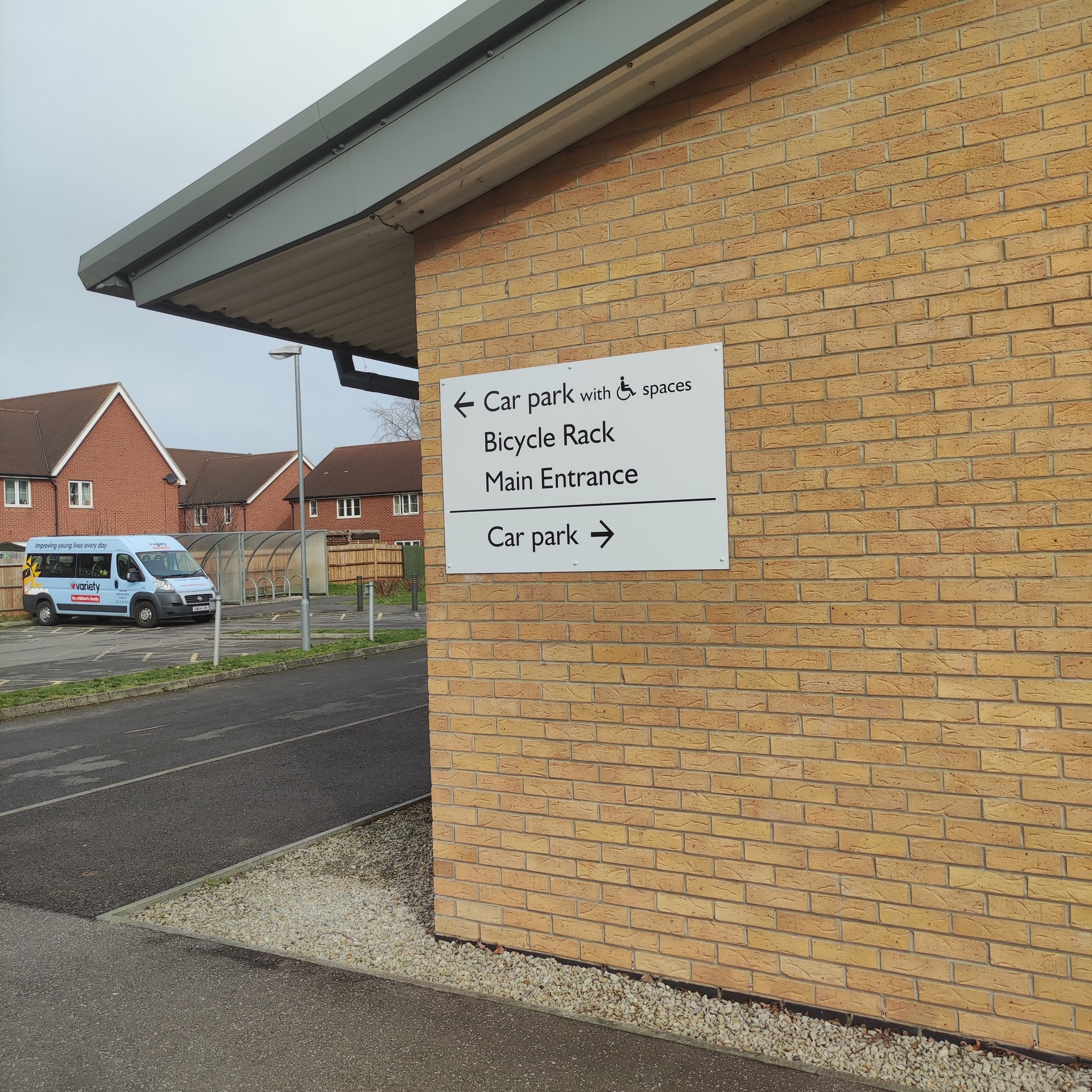 Exterior directional wayfinding sign mounted on a brick wall indicating car park, bicycle rack and main entrance, with arrows and accessible icon.