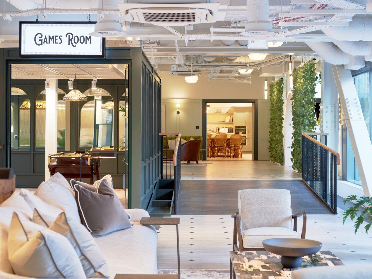 Modern office interior showing a hanging 'Games Room' wayfinding sign above a lounge area with seating, natural light and decorative greenery.
