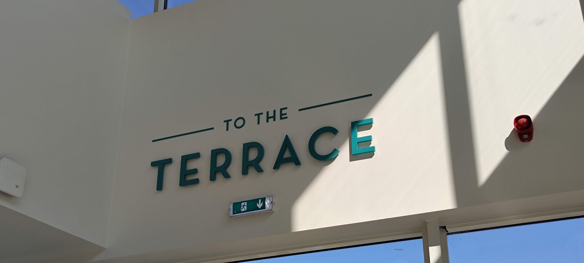 Interior directional sign with raised green letters reading 'TO THE TERRACE' mounted on a pale wall, photographed with strong natural light and shadows.