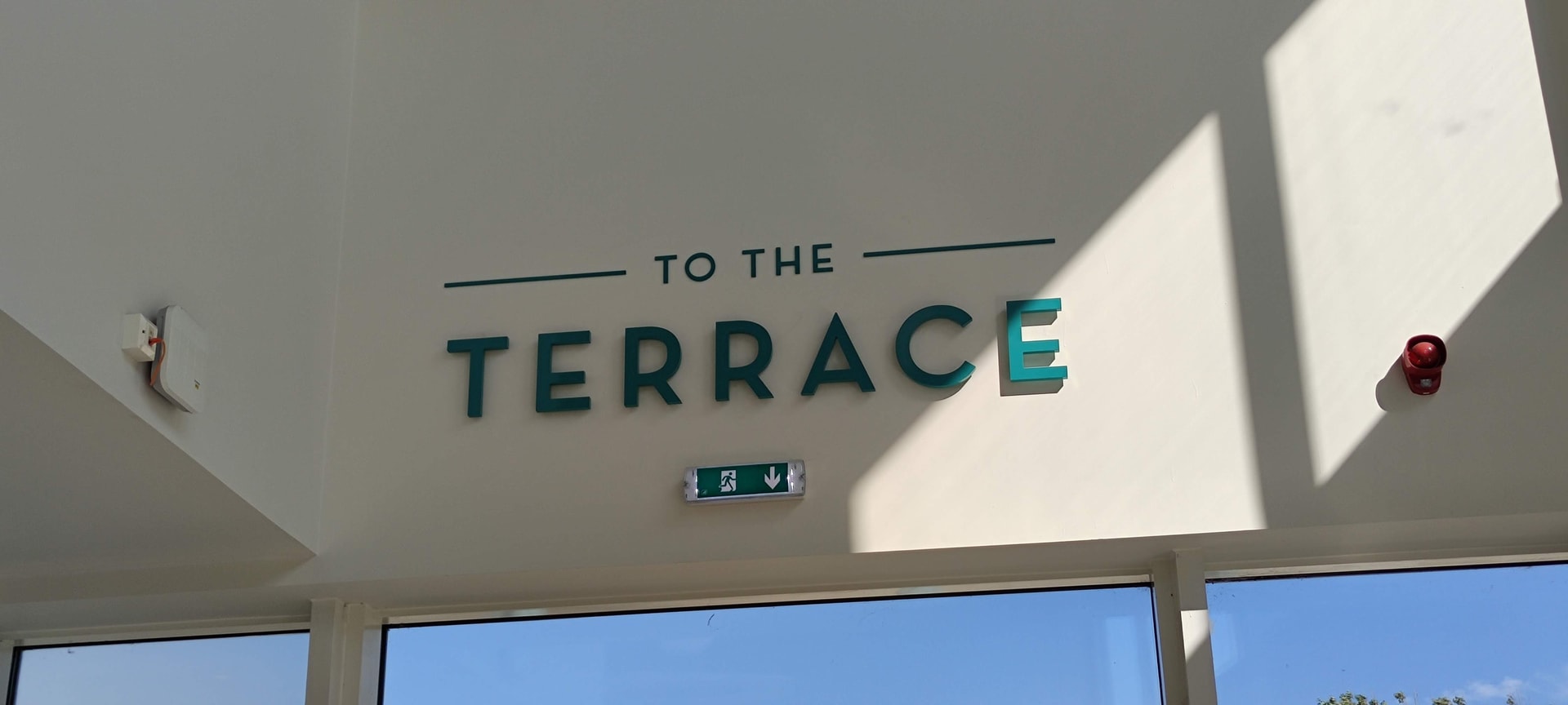 Interior wayfinding sign 'TO THE TERRACE' made from raised teal letters mounted on a white wall above windows, illuminated by natural daylight.