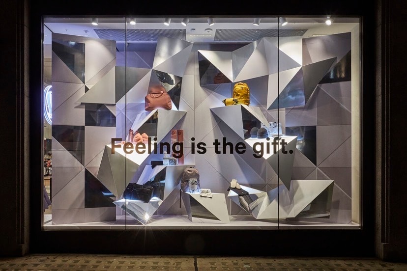 Retail store window display with geometric folded-panel backdrop and vinyl slogan on glass, showcasing bags and shoes under spot lighting.