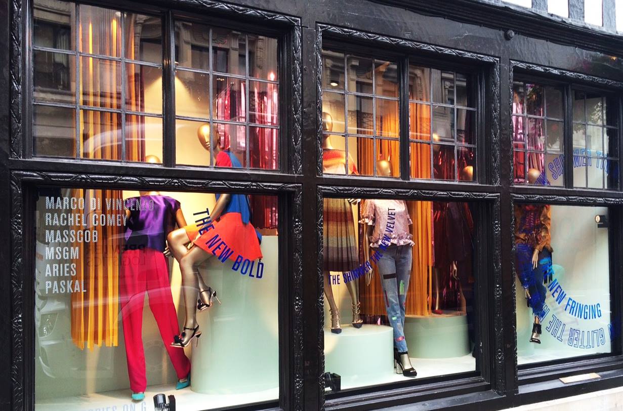 Fashion retail window display featuring mannequins and blue vinyl graphics with designer names and promotional slogans on glass.