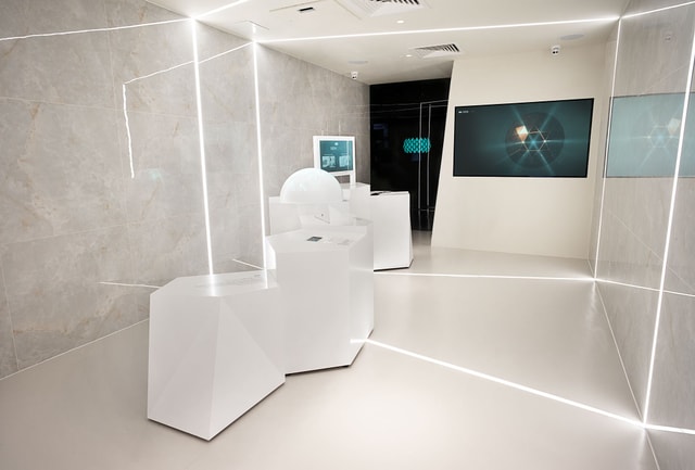 Minimalist exhibition space with geometric white display plinths, integrated LED strip lighting and a large wall-mounted digital screen, showcasing a polished experiential installation.