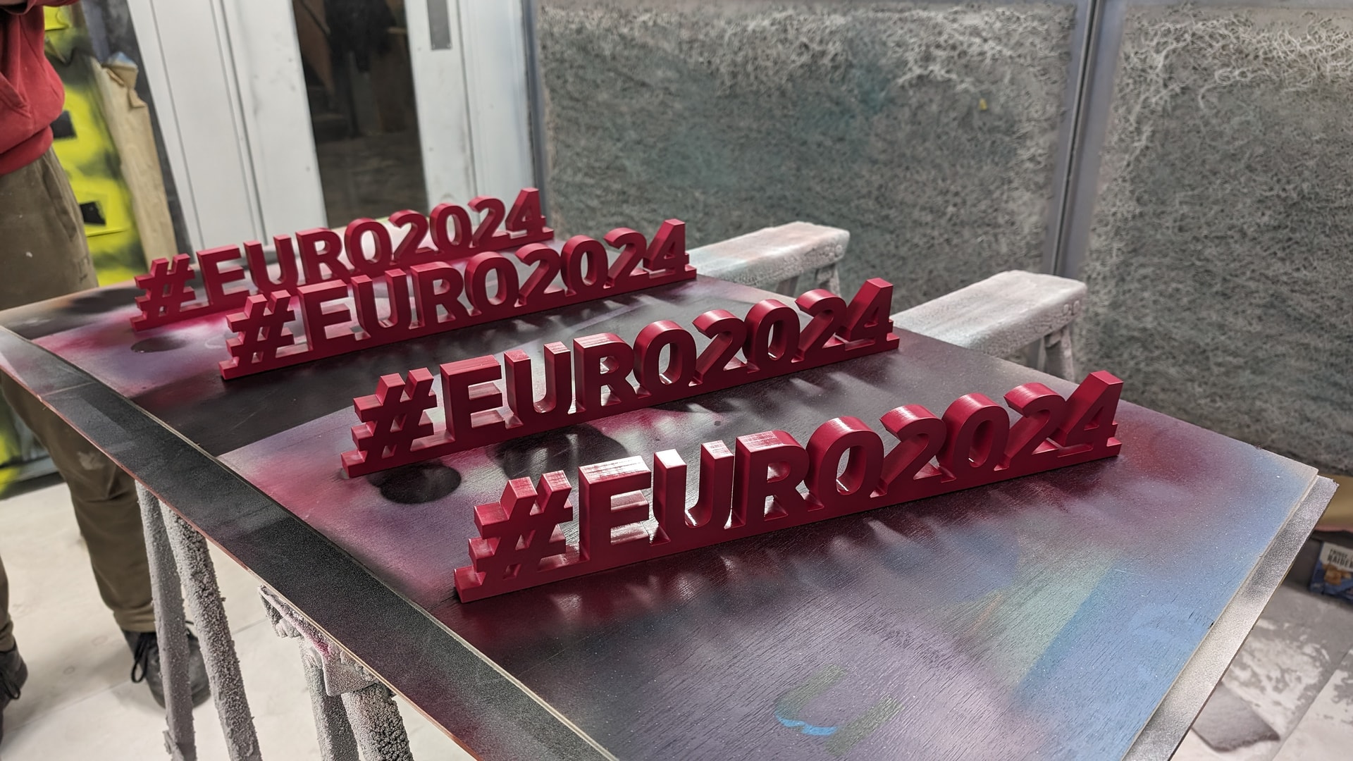 Freshly spray-painted red 3D '#EURO2024' letters drying on a workboard inside a paint booth, showing the finishing stage of event signage production.