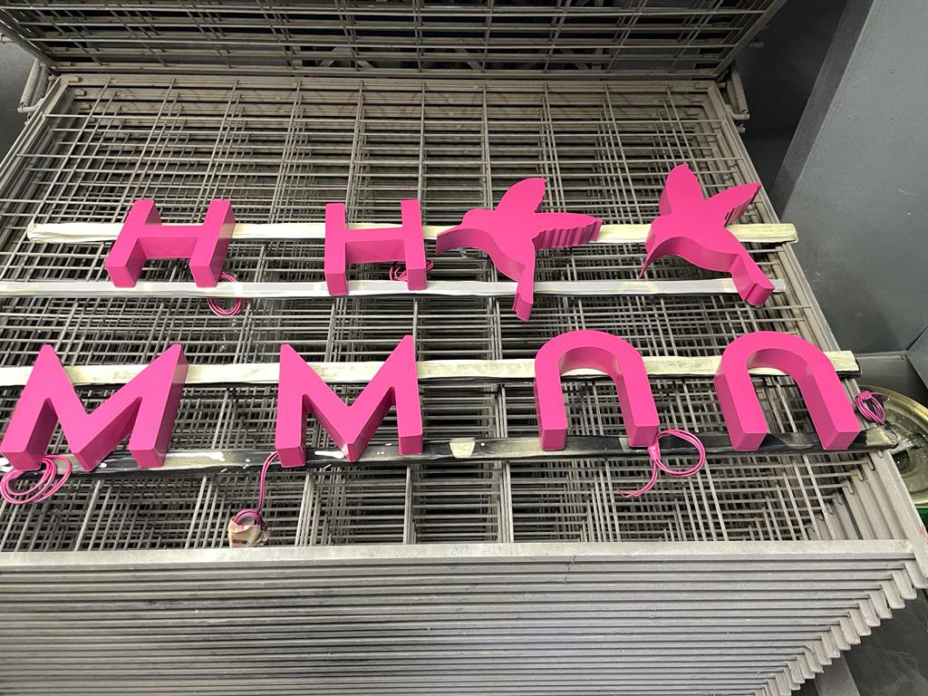 Pink fabricated 3D sign letters with LED wiring drying on a paint/drying rack inside a workshop spray booth.
