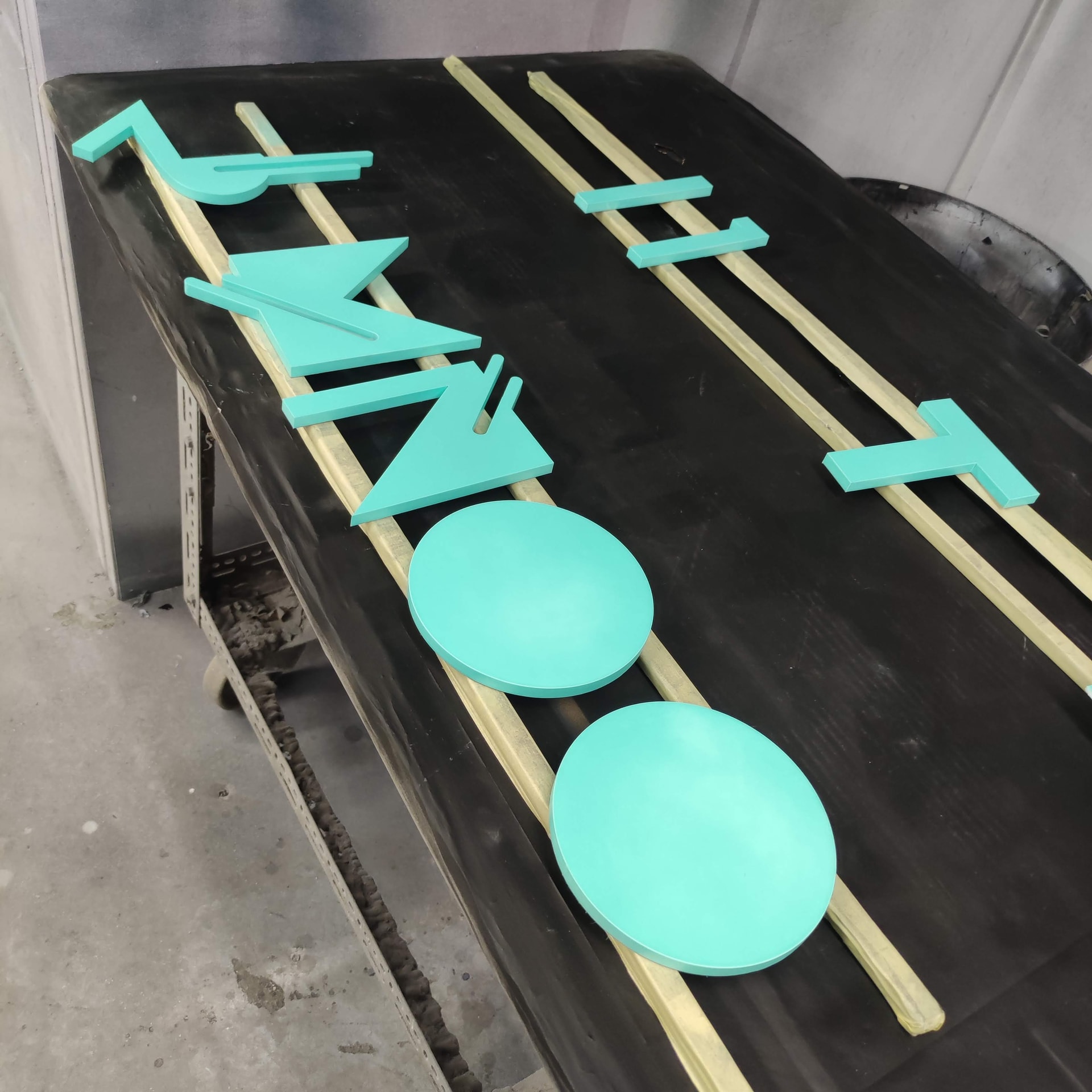 Turquoise acrylic sign letters and round sign elements laid out on a spray table in a finishing booth, showing freshly painted signage components ready for assembly or installation.