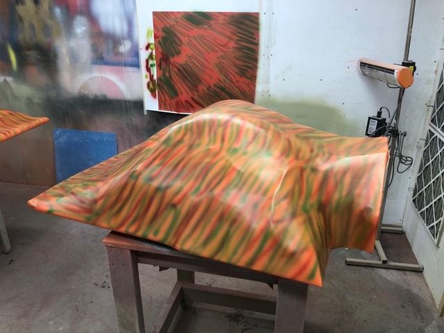 Vacuum-formed plastic shell with a multicolour spray paint finish in a spray booth, shown on a workstand with heater and test panel.