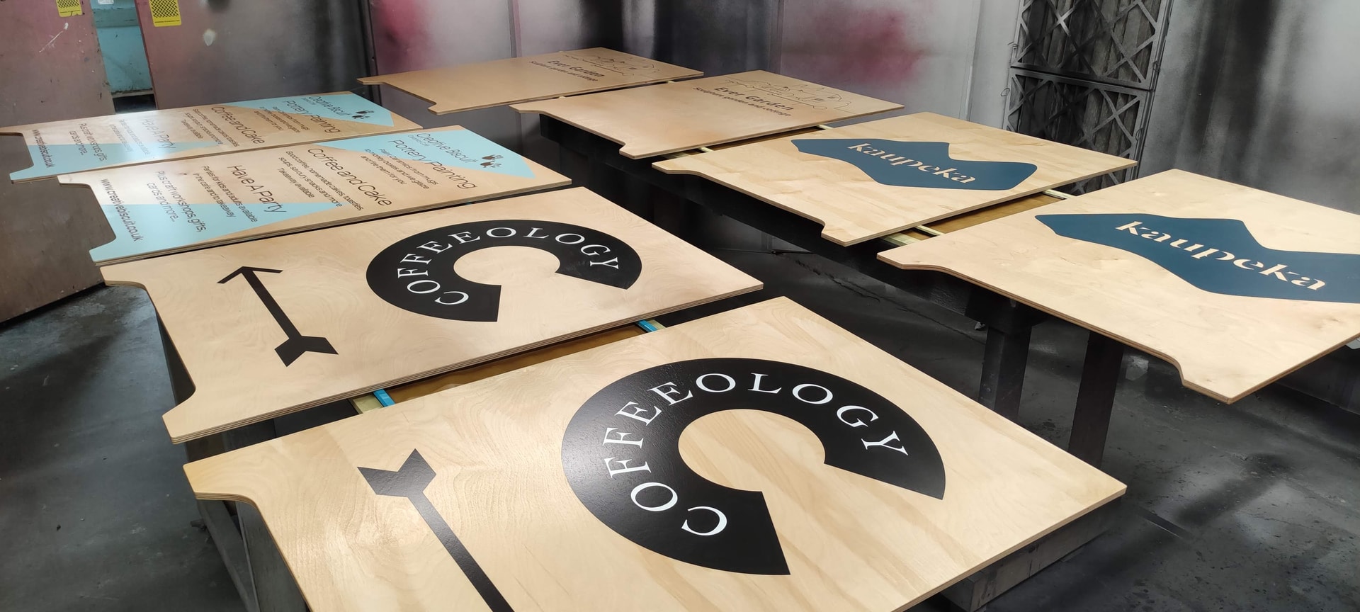 Wooden branded sign panels with Coffeeology logos and directional arrows drying in a spray booth during finishing and painting.