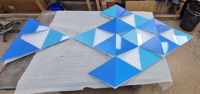 Glossy blue triangular acrylic panels arranged on a workshop bench, staged for finishing and installation.