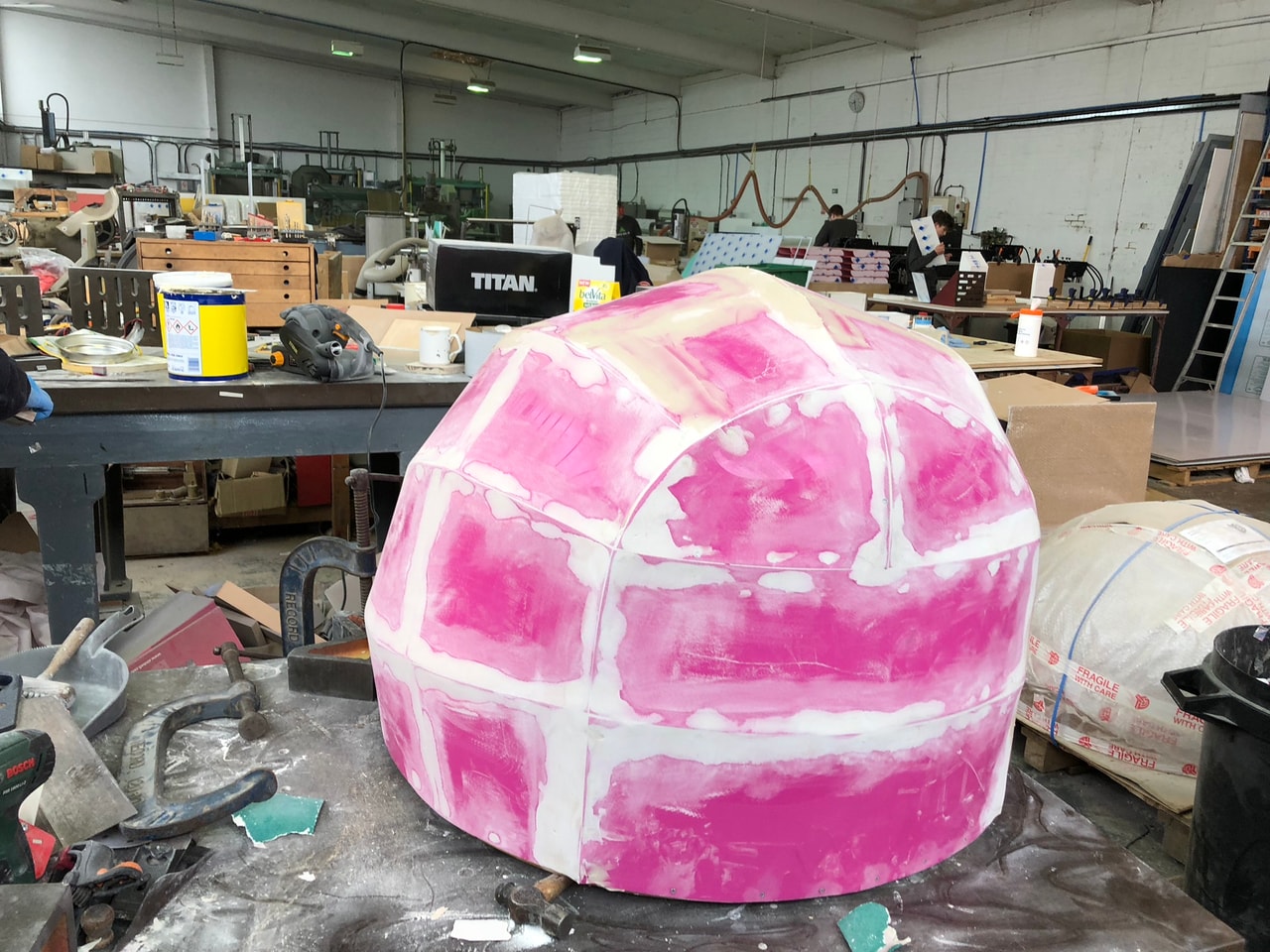 Workshop shot of a large formed shell/dome with pink filler and sanding in progress on a fabrication bench, showing an in-progress plastic/composite fabrication and finishing process.