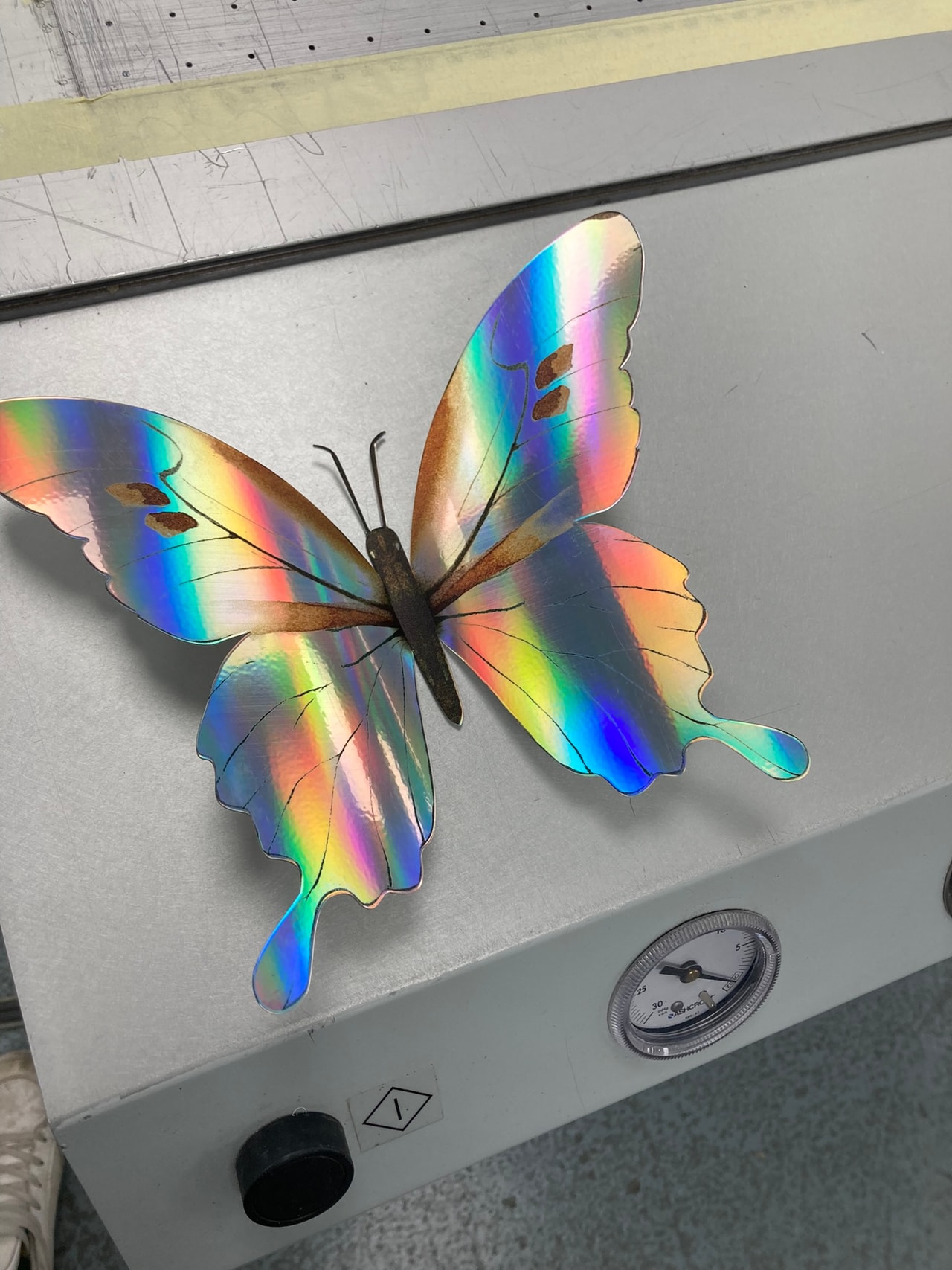 Iridescent holographic butterfly decorative panel on a workshop bench with a pressure gauge and control knob, showing a finished fabricated piece in a production environment.