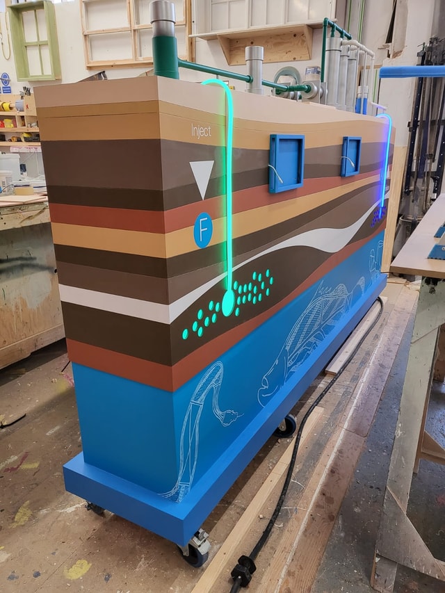 Painted display/fixture with layered graphics and integrated LED tubing on castors photographed in a fabrication workshop.