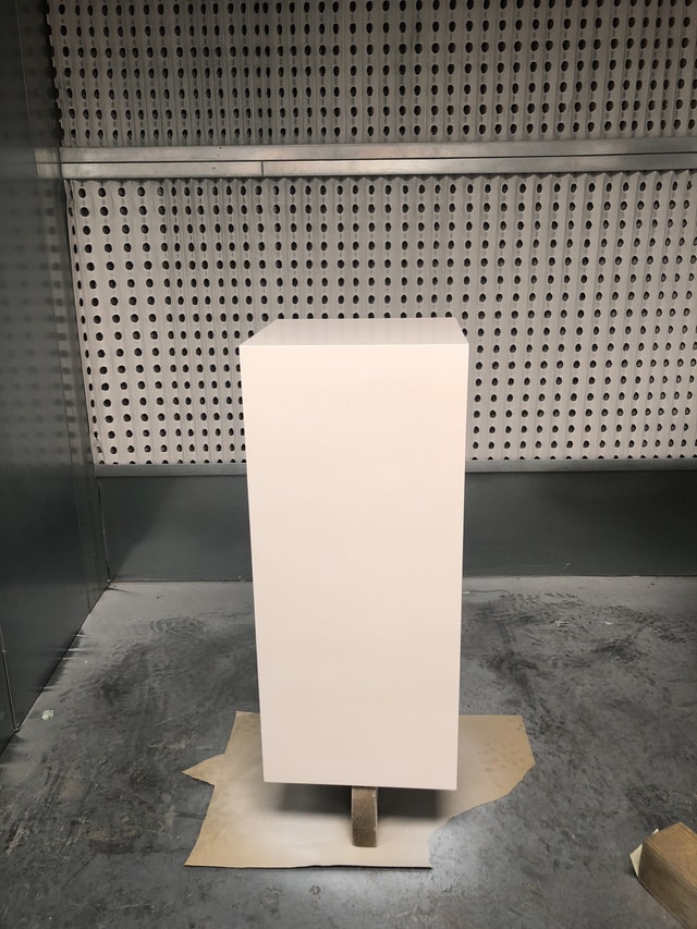 White painted display pedestal photographed in a spray booth, showing a finished/near-finished retail fixture in a fabrication workshop.