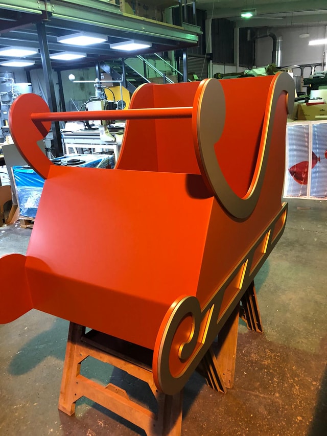 Large red and gold painted sleigh prop on sawhorses in a fabrication workshop, shown as a finished display/props piece ready for events or retail.