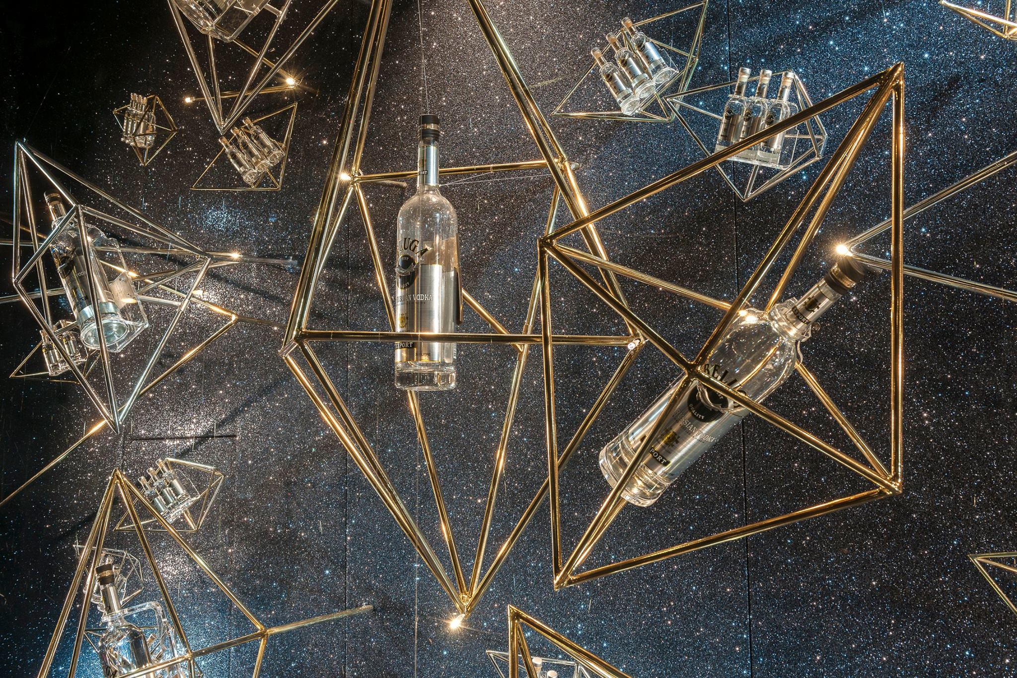 Stylish retail window display featuring suspended geometric brass frames holding glass bottles against a glittering backdrop with integrated lighting.