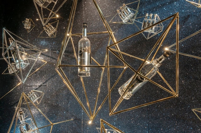 Stylish retail window display featuring suspended geometric brass frames holding glass bottles against a glittering backdrop with integrated lighting.