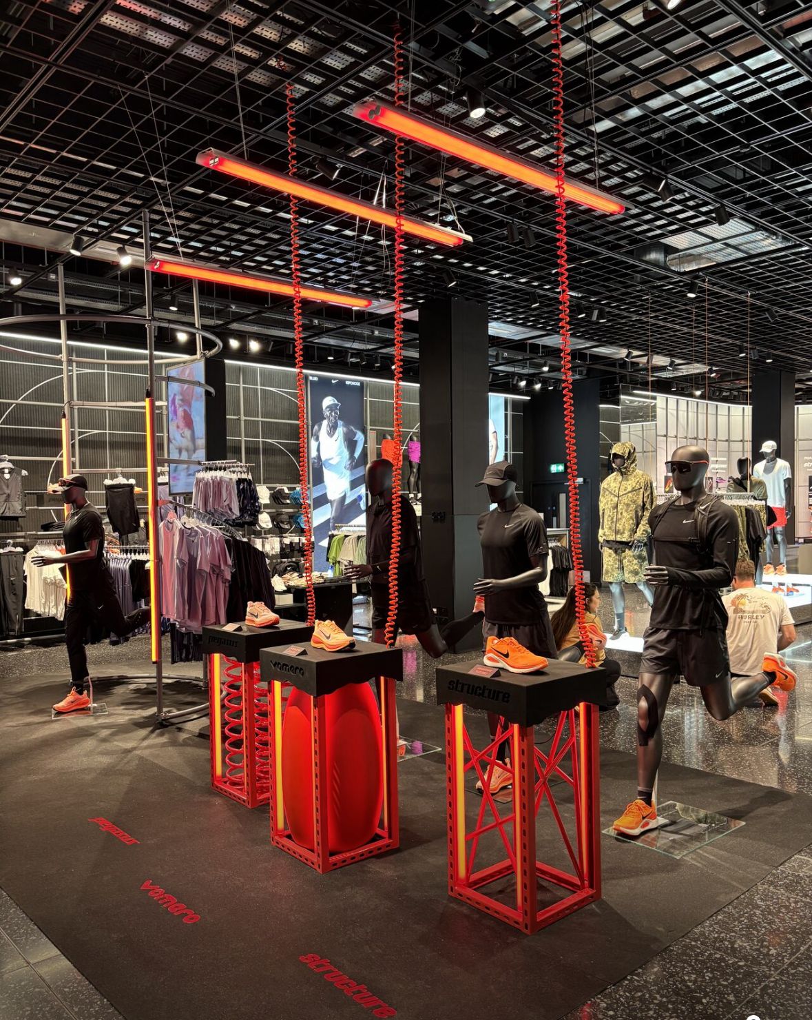 Nike retail store interior showing Vomero sneaker displays on illuminated pedestals, mannequins in athletic apparel and prominent red linear lighting.