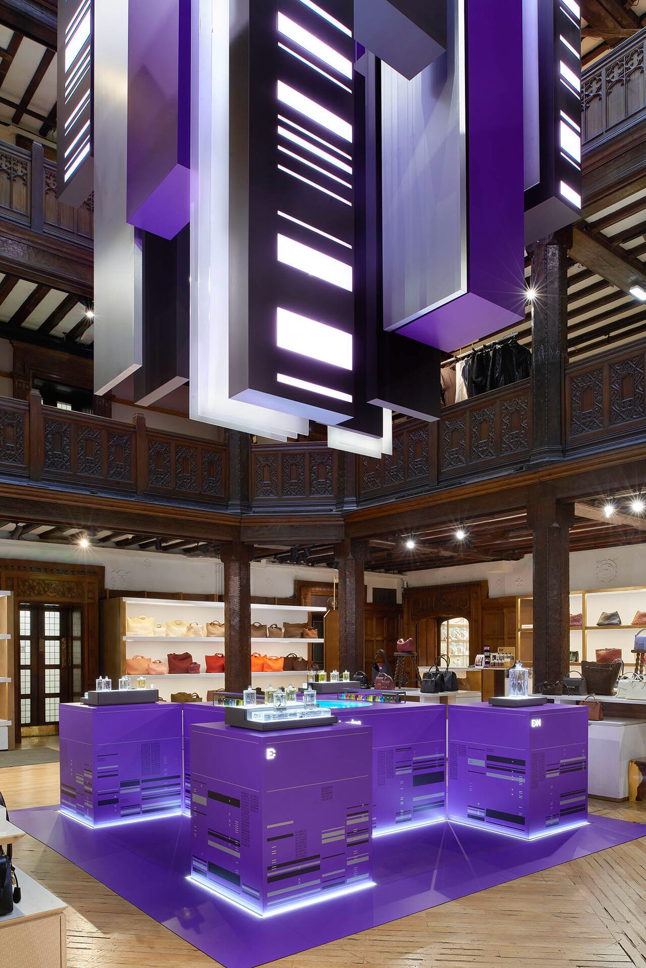 High-end retail interior with purple illuminated display islands and suspended backlit light sculptures showcasing handbags and accessories in a wooden boutique setting.